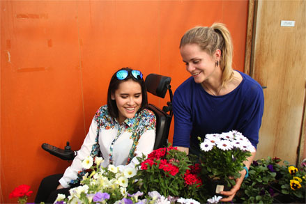 Bilde av en kvinne i rullestol som kikker på blomster. En annen kvinner viser fram ulike blomster i hvitt, rødt, gult og lilla.