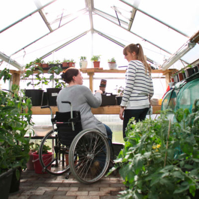 Bilde av to kvinner i et drivhus omkranset av grønne planter. En kvinne sitter foran et bord i rullestol mens den andre kvinnen står oppreist.