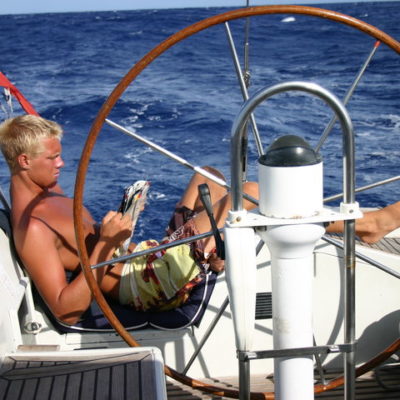 Bilde av artikkelskribent som sitter lettere henslengt akterut i en seilbåt. Han leser i et blad mens han ser ut til å sole seg. I bakgrunnen er det kun hav og i forgrunnen er seilbåtens ror.