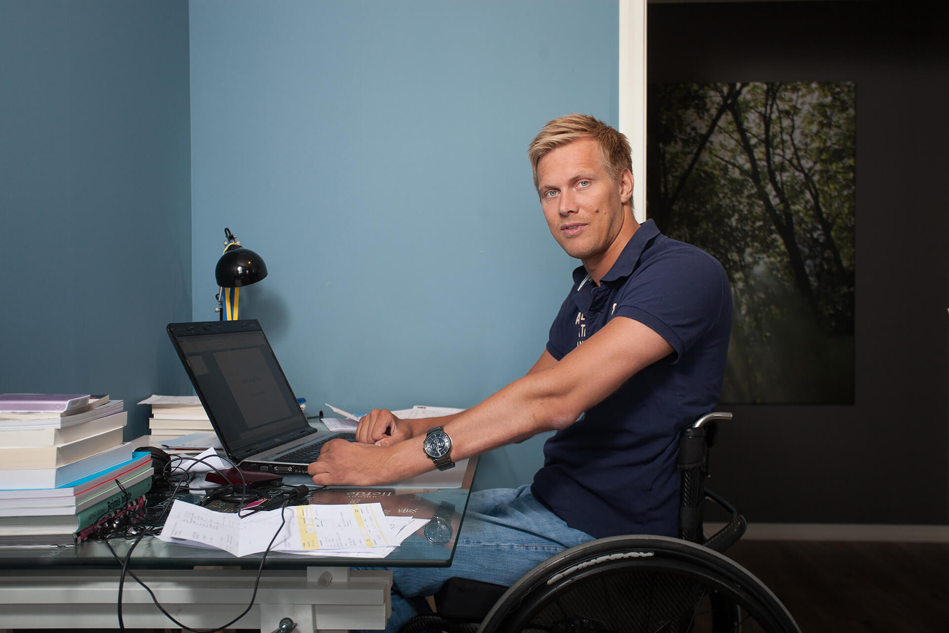Bilde av artikkelskribent som sitter og jobber foran en laptop i rullestol. Han lener armene mot skrivebordet mens han kikker mot kameraet.