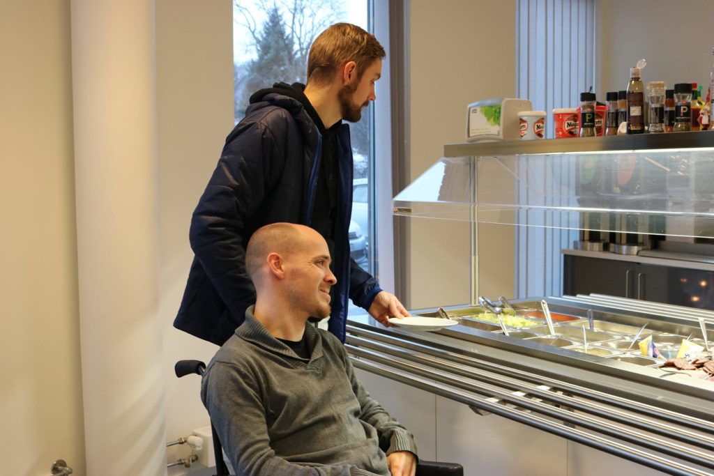 Bilde som viser Kristoffer Svendsen og hans funksjonsassistent, Abraham Heggvik, i kantinen på Kristoffers arbeidsplass. Kristoffer sitter i rullestol ved siden av salatbaren og kikker bort på utvalget. Hans funksjonsassistent står bak ham og holder en tallerken i hånden.