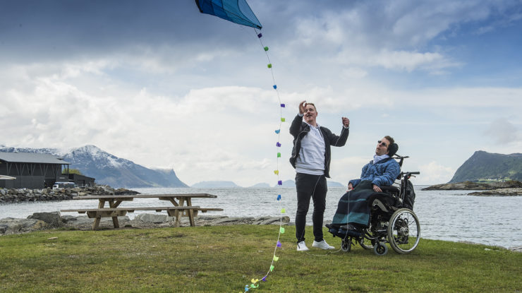 Mannlig assistent og mann i elektrisk rullestol. Assistenten holder i en drage som flyr i vinden. De befinner seg ved sjøen på en grønn plen. Vi ser fjell i bakgrunnen.