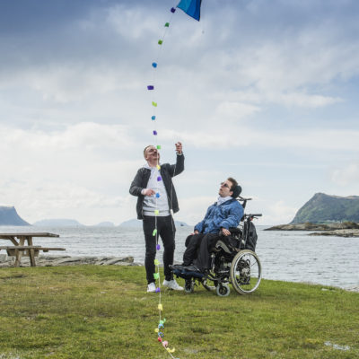 Bilde av mannlig assistent sammen med mann i rullestol. De står sammen på grønt gress foran hav og fjell, hvor assistenten holder i en drage som flyr i luften.