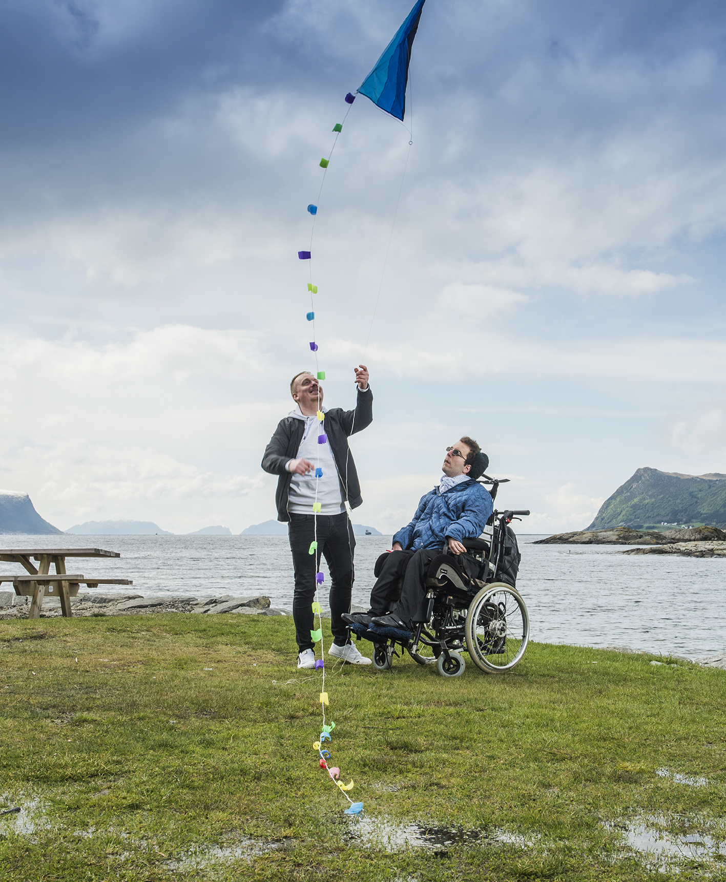 Bilde av mannlig assistent sammen med mann i rullestol. De står sammen på grønt gress foran hav og fjell, hvor assistenten holder i en drage som flyr i luften.