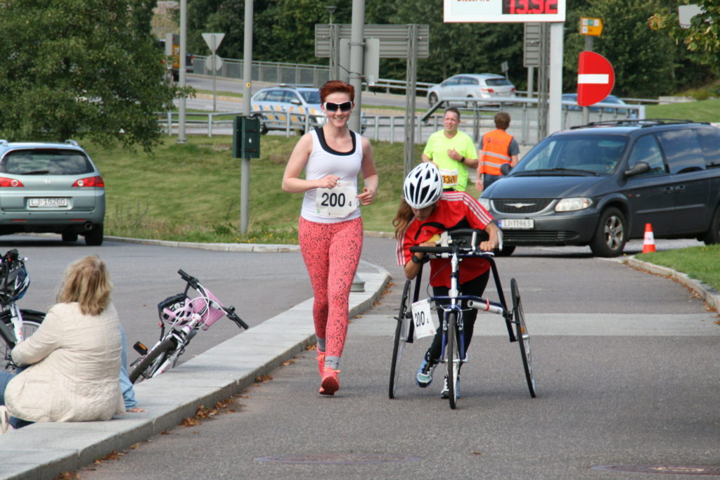 Bilde av parautøver Marte Åsvang med løpesykkel og en annen kvinne uten løpesykkel. De deltar i et RaceRunning-løp og løper begge nedover en vei. I bakgrunnen skimtes en bil og frivillige.