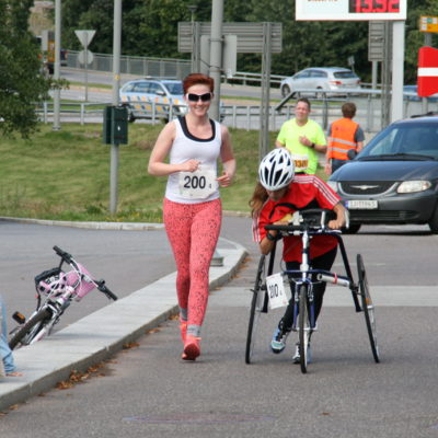 Bilde av parautøver Marte Åsvang med løpesykkel og en annen kvinne uten løpesykkel. De deltar i et RaceRunning-løp og løper begge nedover en vei. I bakgrunnen skimtes en bil og frivillige.