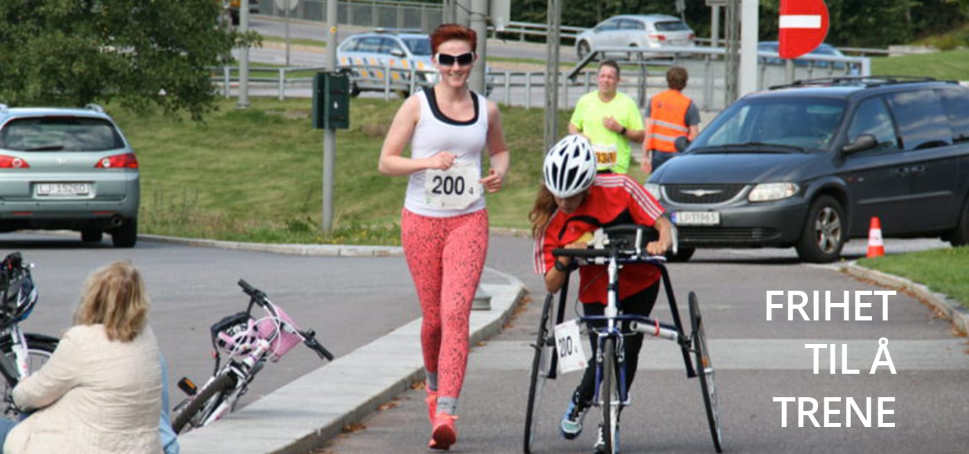 Bilde av parautøver Marte Åsvang med løpesykkel og en annen kvinne uten løpesykkel. De deltar i et RaceRunning-løp og løper begge nedover en vei. I bakgrunnen skimtes en bil og frivillige. 