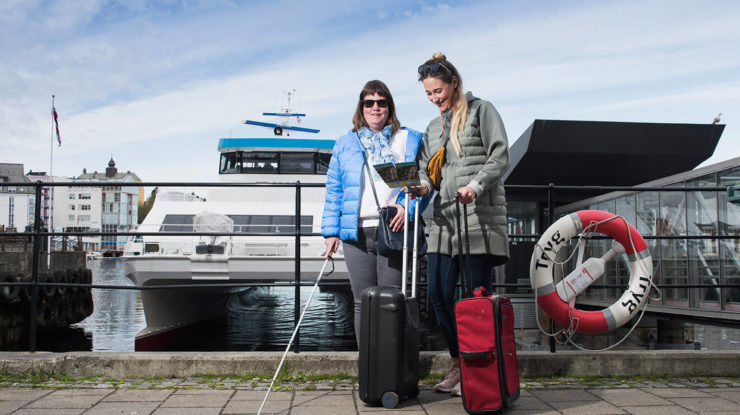 Kvinnelig assistent bistår synshemmet kvinne på reise. De har to kofferter foran seg og står på fergekaien foran en båt.