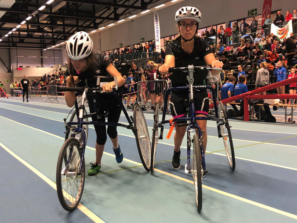 Bilde av artikkelskribent Sofie Østerbø Jansen og en annen utøver. De er begge på banen for å gjennomføre et løp i Racerunning. Sofie kikker inn i kamera mens hun holder rundt løpesykkelen.