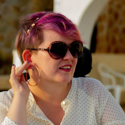 Nærbilde av Kine Lyngås. Hun kikker til siden mens hun berører en ørering med den ene hånden. Hun har på seg en prikkete skjorte og solbriller.