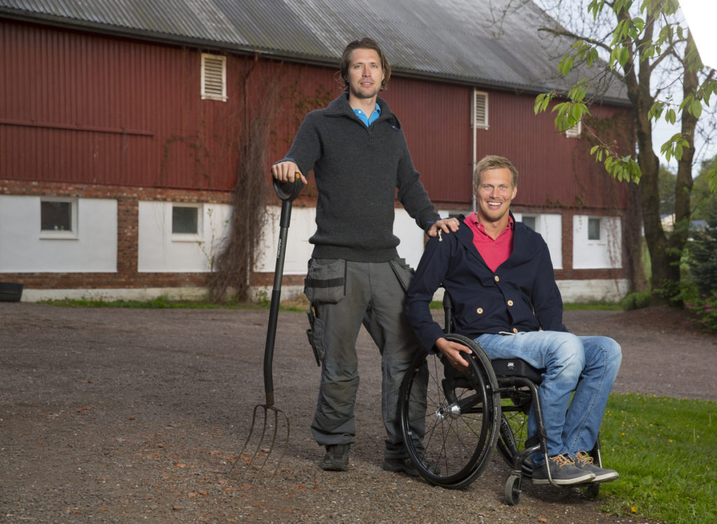 Bilde av Lars Ivar Eilerås, markedskoordinator i Prima Assistanse. Han sitter i rullestol og kikker smilende mot kameraet. Ved siden av seg står hans bror med en høygaffel i hånden. I bakgrunnen skimtes et gårdsbygg.