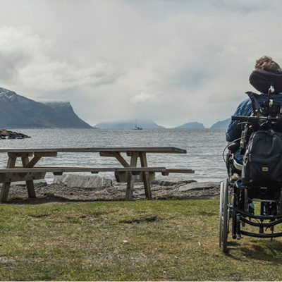 Bilde av en kvinne i rullestol med en mann som sitter på kne ved siden av henne. De har begge ryggen mot kameraet mens de kikker ut mot en fjord. Foran dem er et rasteplassbord.
