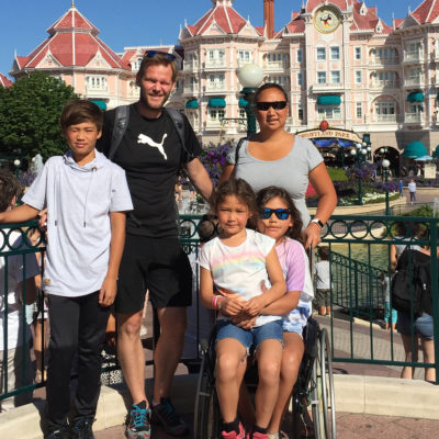 Bilde av BPA-koordinator Elisabeth Lillegrend sammen med sin ektemann, sønn og to døtre. Den ene datteren sitter i rullestol med sin søster på fanget. De andre står bak dem. I bakgrunnen skimtes et bygg med påskriften Disneyland.