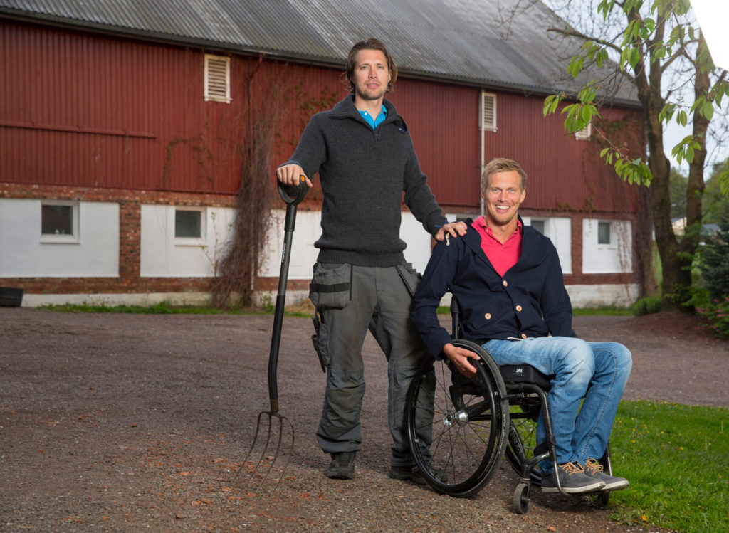 Bilde av Lars Ivar Eilerås, markedskoordinator i Prima Assistanse. Han sitter i rullestol og kikker smilende mot kameraet. Ved siden av seg står hans bror med en høygaffel i hånden. I bakgrunnen skimtes et gårdsbygg.