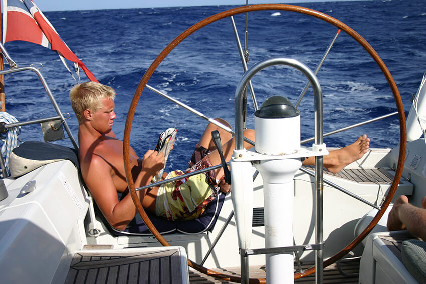 Bilde av artikkelskribent som sitter lettere henslengt akterut i en seilbåt. Han leser i et blad mens han ser ut til å sole seg. I bakgrunnen er det kun hav og i forgrunnen er seilbåtens ror. 