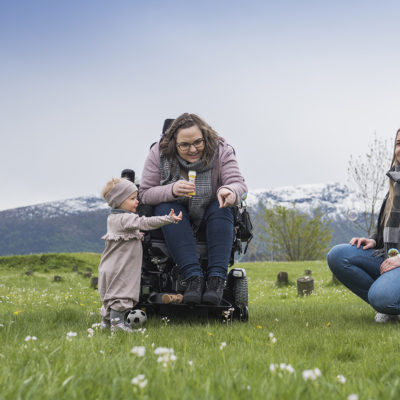 Kvinne i rullestol sammen med det lille jentebarnet sitt på grønt gress og bed fjell i bakgrunnen. Kvinnen peker på noe. Kvinnelig assistent sitter ved siden av og smiler.