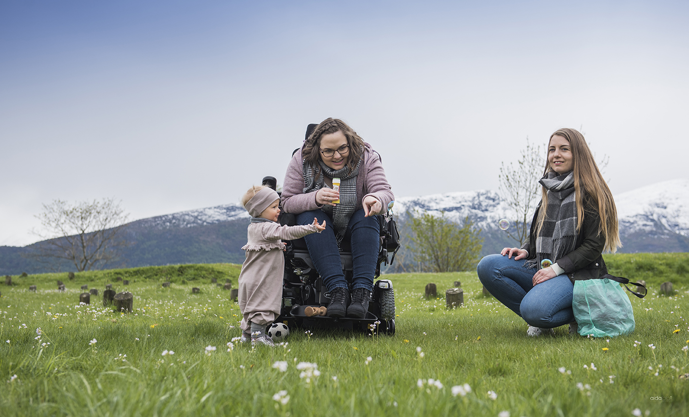 Kvinne i rullestol sammen med det lille jentebarnet sitt på grønt gress og bed fjell i bakgrunnen. Kvinnen peker på noe. Kvinnelig assistent sitter ved siden av og smiler.