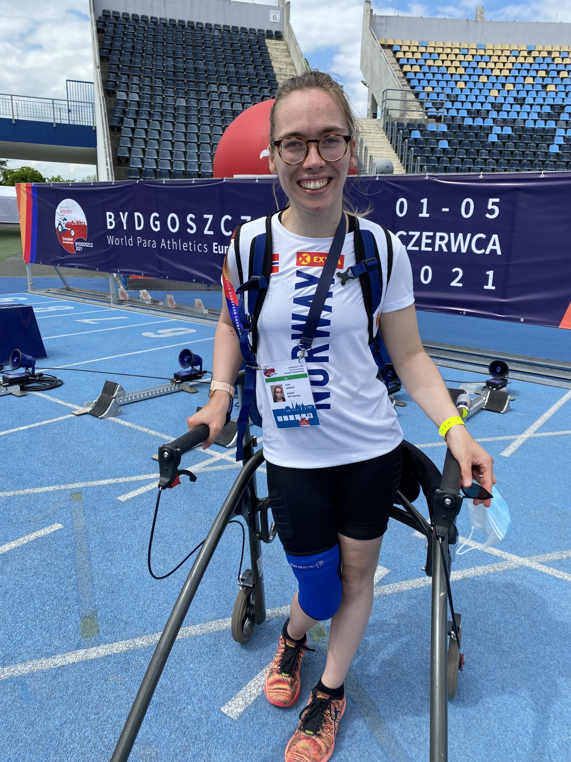 Sofie smiler til kamera fra idrettsanlegg. Hun står med en rullator.