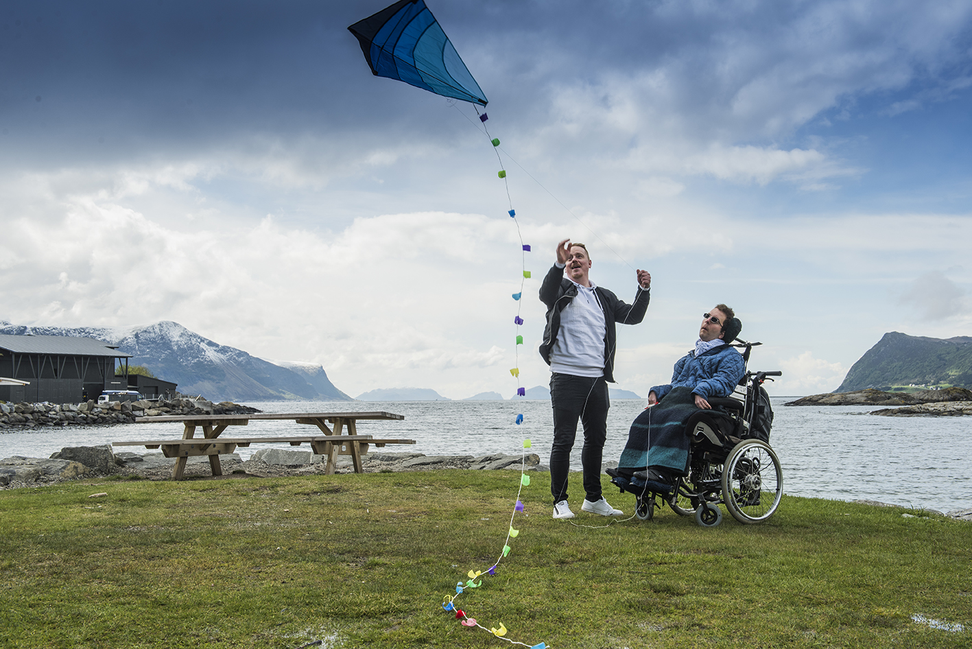 Mannlig assistent hjelper mann i rullestol med å fly en drage. I bakgrunnen ser man flotte fjell og sjø.