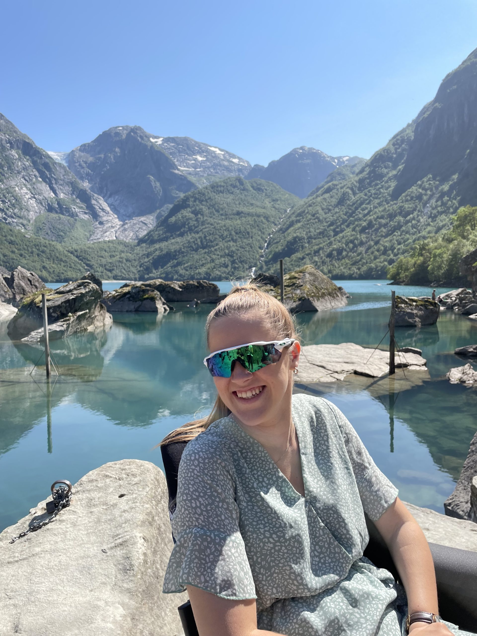 Hannah har på raske skibriller og sitter i en grønngrå kjole i rullestolen sin foran en innsjø med fjelltopper i bakgrunnen. Smiler stort og det er en solfylt dag.