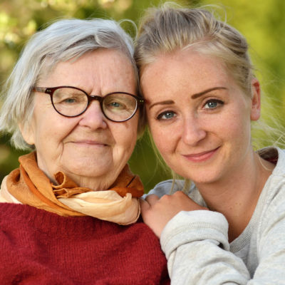 Eldre dame sammen med kvinnelig assistent som holder armen rund henne. De smiler begge til kamera.