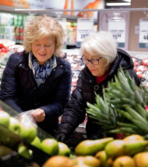 Kvinnelig assistent bistår eldre dame på dagligvarebutikken. De befinner seg i fruktavdelingen.