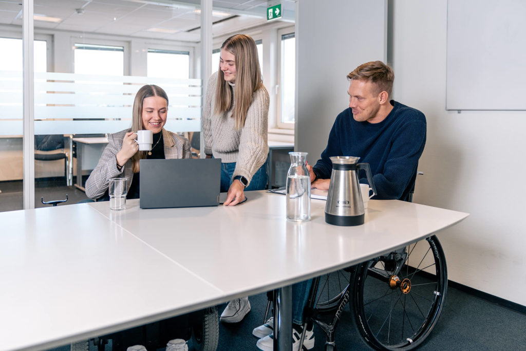 Kollegaer Hannah og Lars, begge i rullestol, på møterom på kontoret. Hannah i beige dressjakke og svart genser. Hun drikker kaffe og smiler. Lars i mørkeblå genser smiler. Assistent i beige genser retter på Hannahs PC.