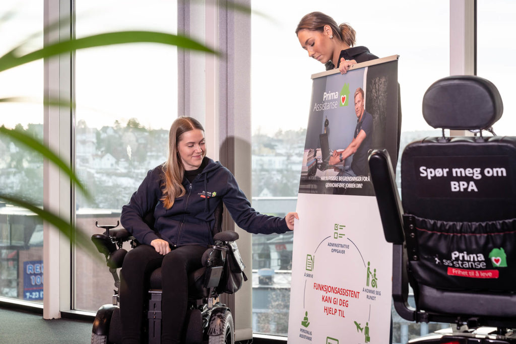 Hannah foran store vinduer sammen med funksjonsassistent. Assistenten holder en roll-up med info om funksjonassistanse. Kontorlandskap. Hannah smiler og har på seg svart bukse og blå Prima-hoodie.