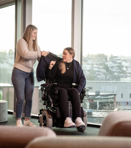 Ung kvinnelig assistent hjelper ung kvinne i elektrisk rullestol med å ta på seg en jakke. Kontorlandskap.