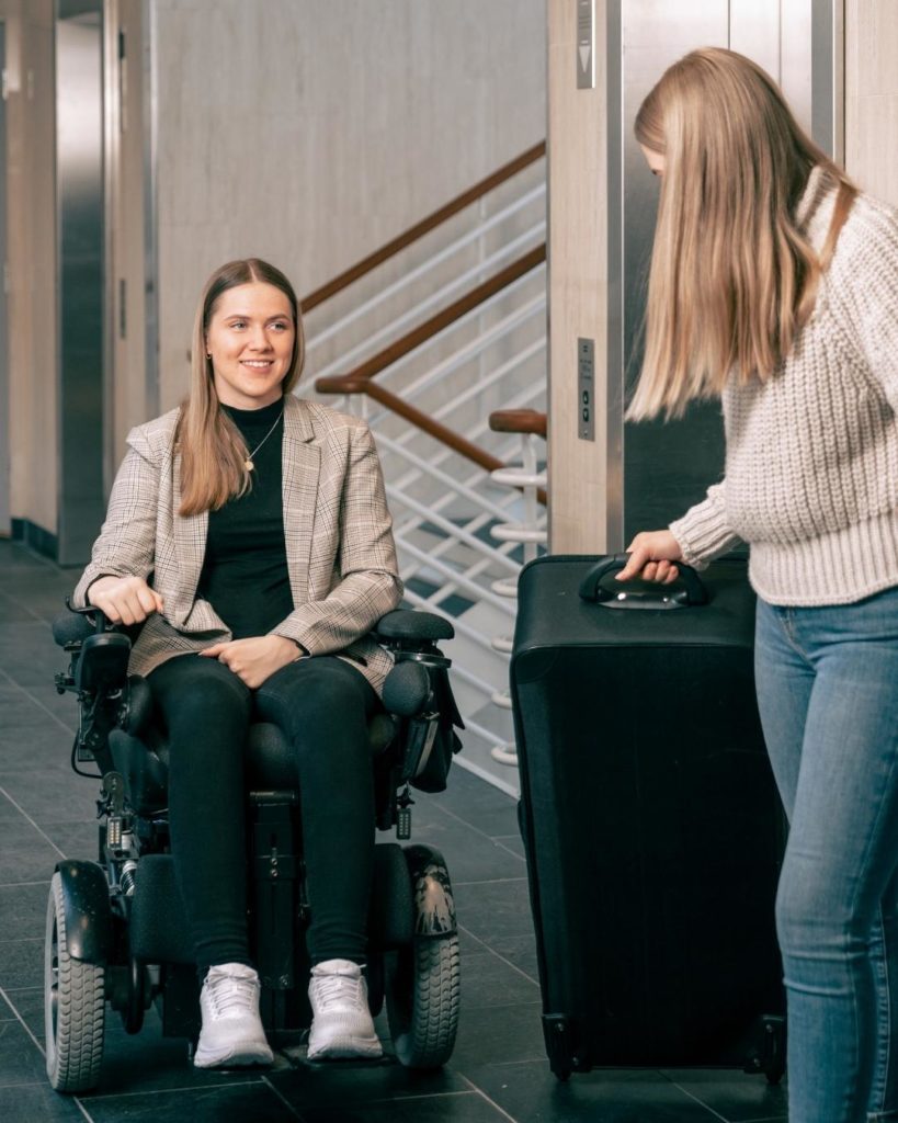 Blond assistent i beige genser og lyseblå jeans, med en svart, stor koffert, åpner døren for Hannah i rullestol. Hannah har på seg beige og rutete dressjakke, svart genser og bukse og hvite sko. Kontorlandskap.