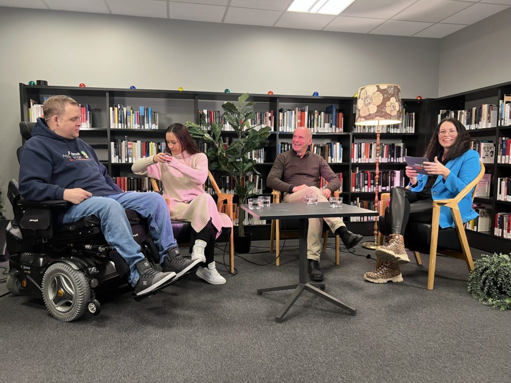 Geir og Hege med to podkastgjester, Lise og Kim, i studio på biblioteket. Bøker og planter i bakgrunnen. God stemning. Lise har på beige strikkegenser, og Kim, som sitter i rullestol, har på blå Prima Assistanse-hettegenser.