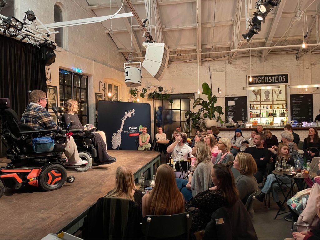 Hannah Lunden og Simen Andersen på scenen på utestedet "Ingensteds" foran et godt publikum. Simen i rutete skjorte og Hannah i svart, glitrende gallakjole. Begge sitter i rullestol. Gammelt fabrikklokale hvor det er veldig høyt under taket.