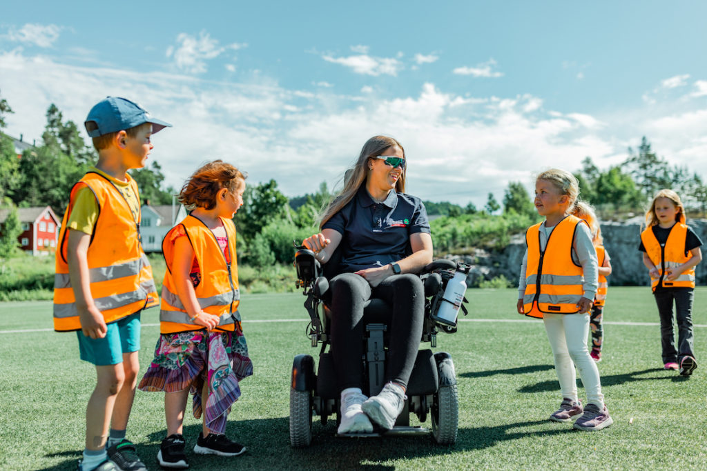 Hannah på fotballbane sammen med en gjeng barn. Hannah har blondt, langt hår og har på seg en blå Prima Assistanse t-skjorte. Hun sitter i elektrisk rullestol. Barna har på oransje refleksvester. Alle smiler. Fint vær.