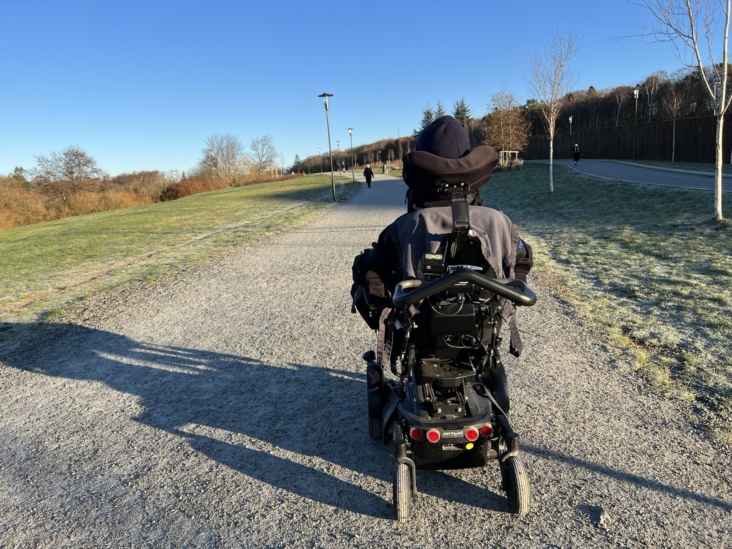 Vi ser Fredrik i sin elektriske rullestol bakfra, mens han kjører på grusstien. Blå himmel og grønt gress.