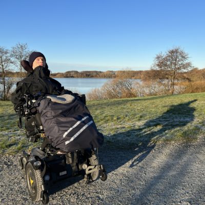 Fredrik sitter godt kledd i elektrisk rullestol foran Mosvannet. Nydelig, blå himmel og grønt gress.
