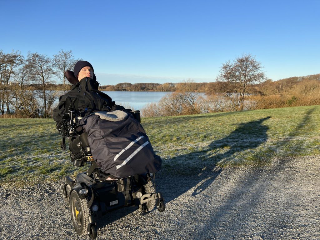 Fredrik sitter godt kledd i elektrisk rullestol foran Mosvannet. Nydelig, blå himmel og grønt gress.