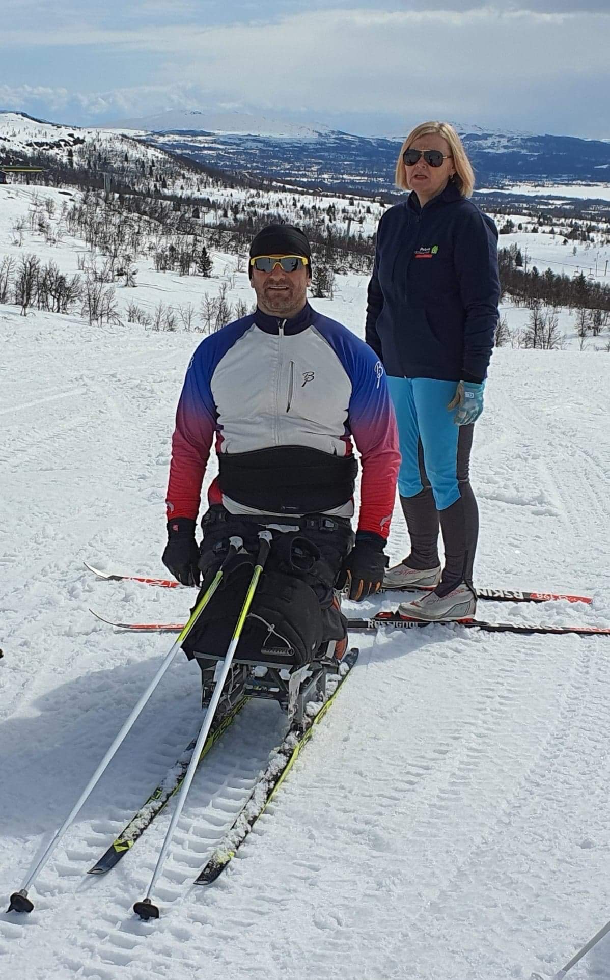 Vigdis og Shaqir på ski. Vigdis har på seg azurblå bukser og mørkeblå hettegenser. Shaqir har på seg en hvit, blå og rød skijakke. Begge har på solbriller. Snøkledde åser i bakgrunnen.