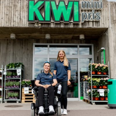 Hannah i elekstrisk rullestol sammen med funksjonsassistent Gina foran Kiwi-butikk. Utenfor butikken er det plassert blomster og planter. Hannah og Gina har på seg mørkeblå t-skjorte med Prima Assistanse-logo og svarte bukser. Begge er blonde og smiler til kamera.