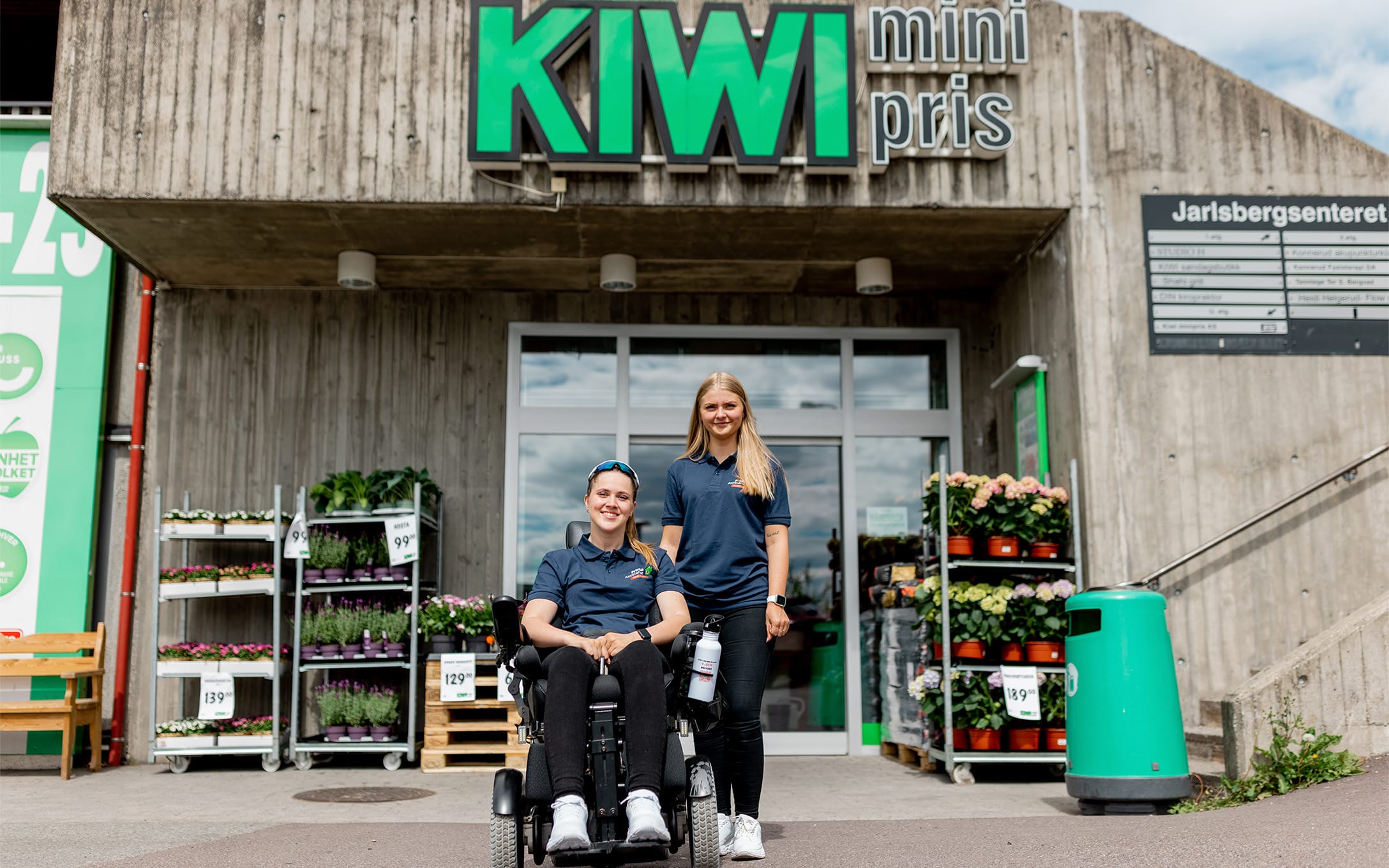 Hannah i elekstrisk rullestol sammen med funksjonsassistent Gina foran Kiwi-butikk. Utenfor butikken er det plassert blomster og planter. Hannah og Gina har på seg mørkeblå t-skjorte med Prima Assistanse-logo og svarte bukser. Begge er blonde og smiler til kamera.