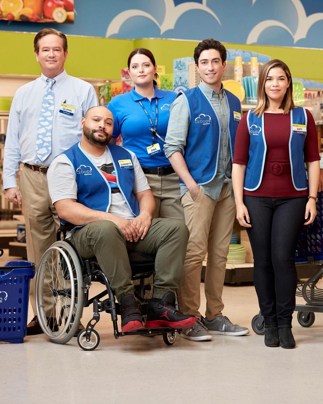 Bildet viser noen av karakterene inne i dagligvarebtuikken i serien "Superstore". 3 menn og 2 damer, kledd i blå vester. De smiler mot kamera. Karakteren Garrett i rullestol fremst i bildet.
