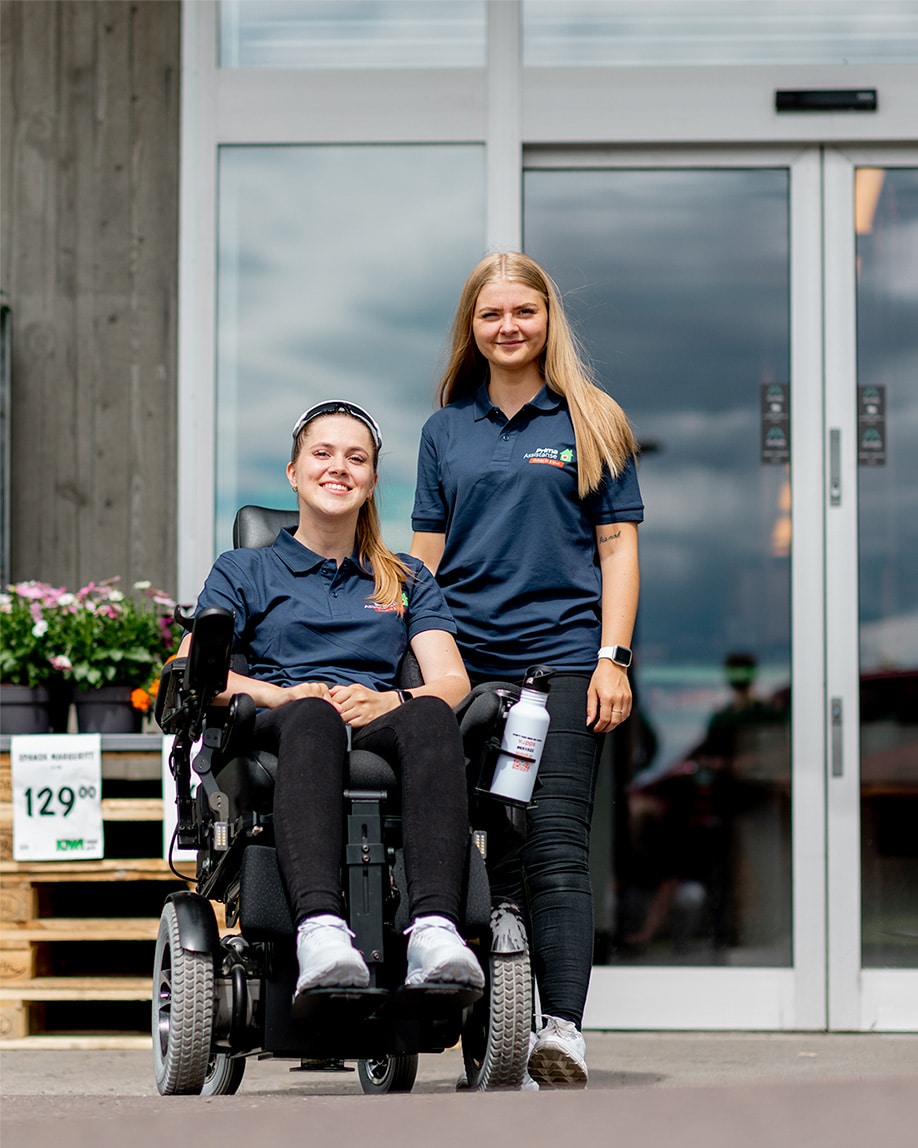 Nærbilde av Hannah i elektrisk rullestol og funksjonsassistent Gina utenfor kiwi-butikk. De smiler. Begge har blondt hår, mørkeblå Prima Assistanse t-skjorter og svarte bukser.