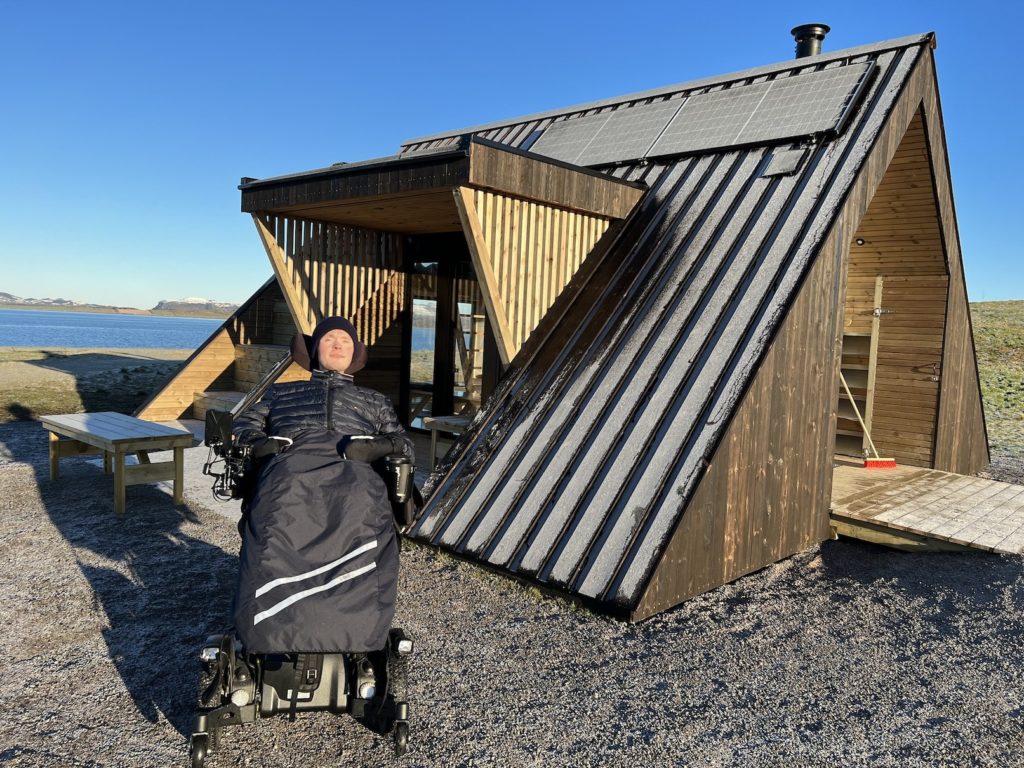 Fredrik i elektrisk rullestol foran Vennskapshyttå. Den er formet som en trekant og er laget av tre, beiset mellommørkt. Man ser hav og gress i bakgrunnen.