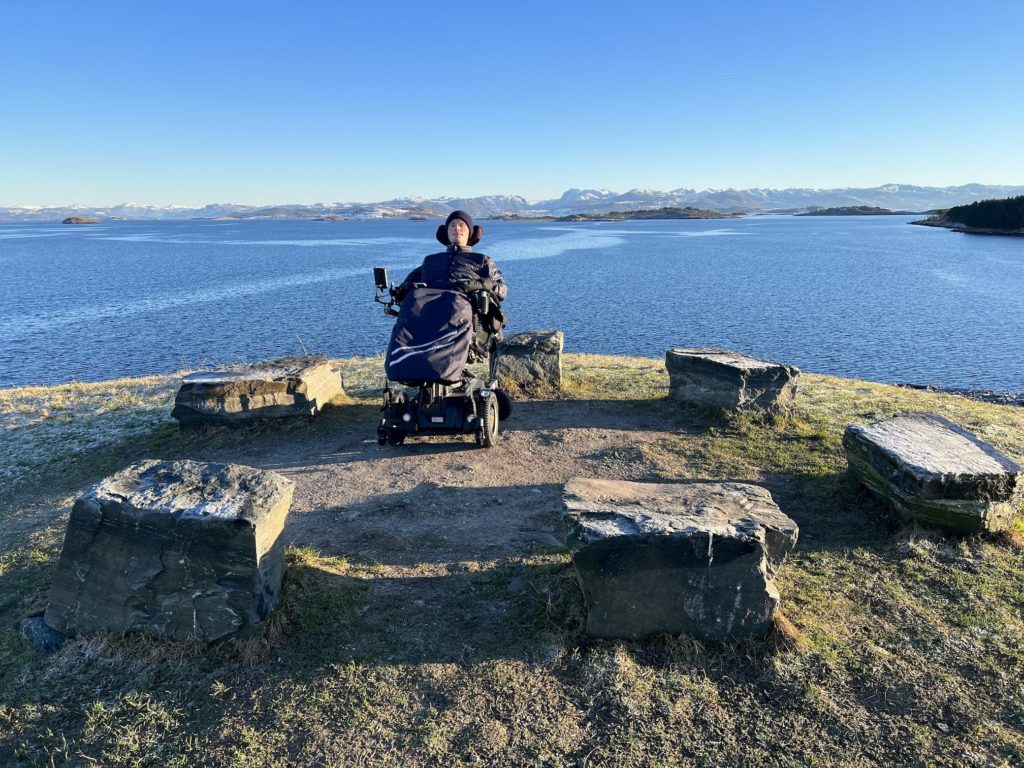 Fredrik på en åstopp med gress i elektrisk rullestol i midten av seks lave steinblokker som former en sirkel. Knallblå himmel og hav i bakgrunnnen, hvor man kan se bevegelse i vannet fra vinden. Fjell i bakgrunnen.