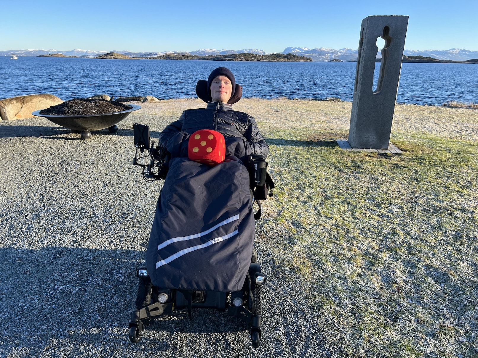 Fredrik i elektrisk rullestol foran minnesmerket, en steinblokk som er hul i midten, for 22. juli på Lundsneset. Flatt terreng, like ved havet. Grønt gress. Han har en stor og rød terning på fanget, som viser terningskast fem.