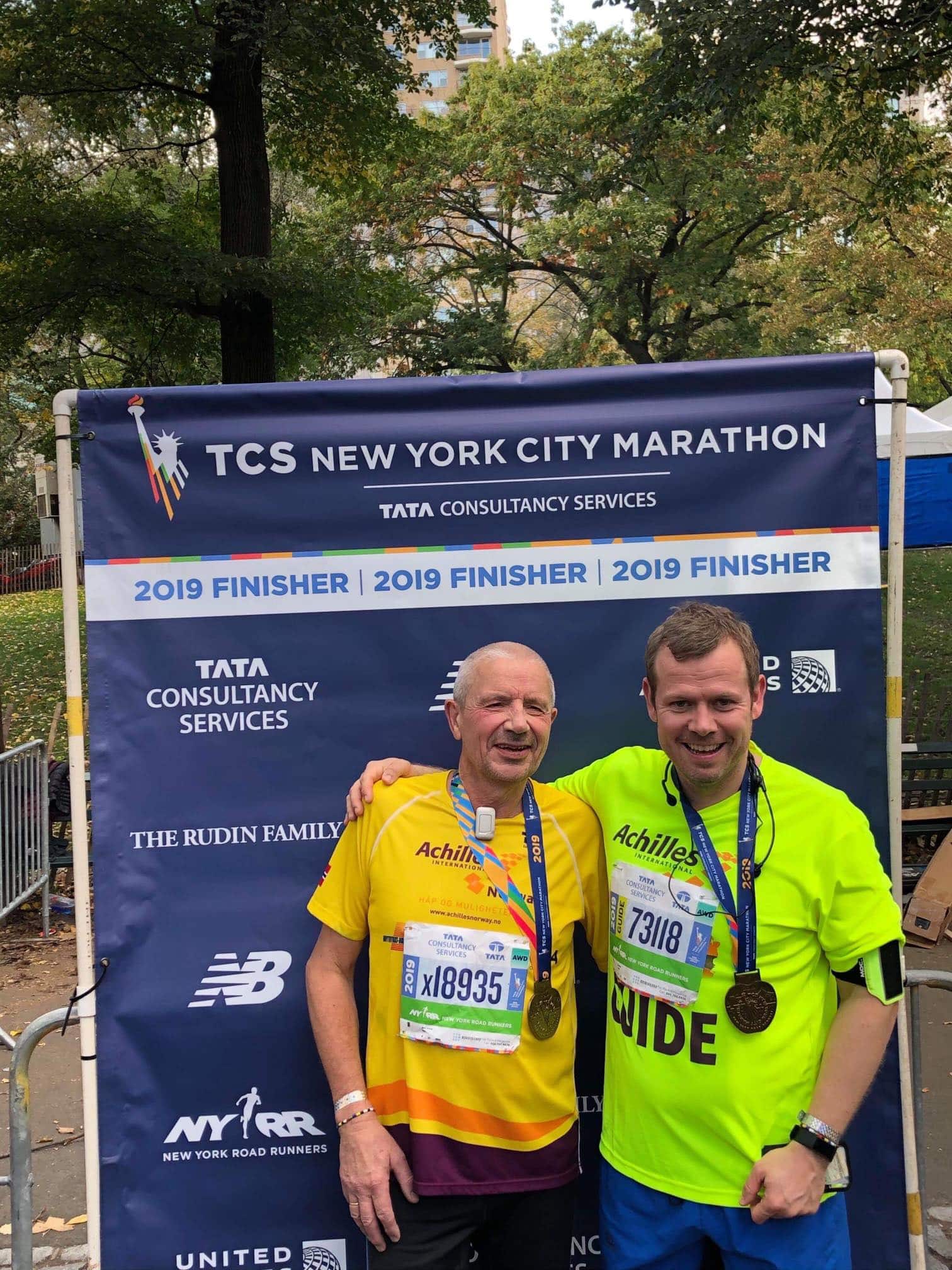 Hans Joakim i gul trøye smiler foran en plakat med New York City Marathon-tekst og arrangementets sponsorer. Assistent holder rundt skulderen hans og smiler. Han har på neongul trøye. Begge har medaljer rundt halsen.