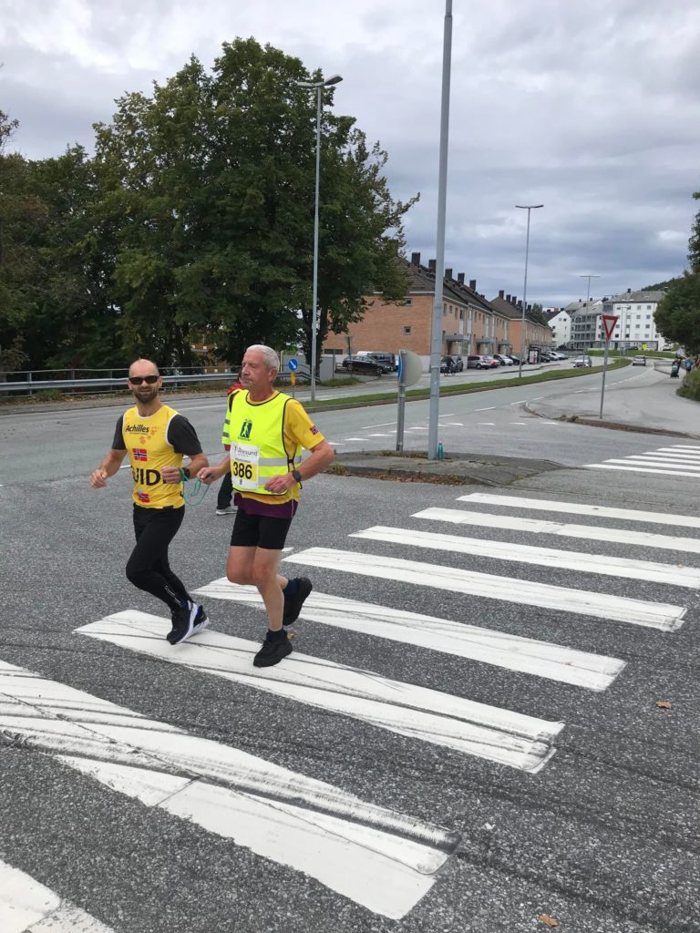 Hans Joakim løper sammen med assistent over fotgjengerovergang. Grønne trær i bakgrunnen. Grå skyer. De er knyttet sammen med en hyssing.