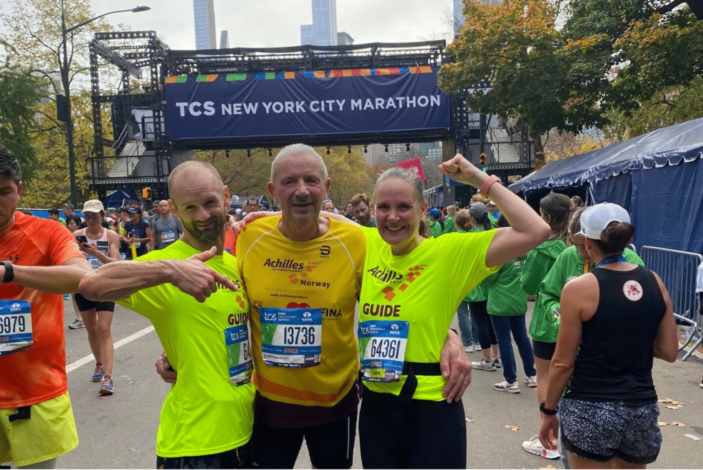 Hans Joakim etter målstreken. Han står i gul trøye og holder rundt sin datter og sin mannlige assistent i neongule trøyer. Bak dem ser man en lilla banner med New York City Marathon skrevet på. Man ser flere løpere stå rundt i bildet, og i bakgrunnen kan man se noen skyskrapere. Alle smiler.