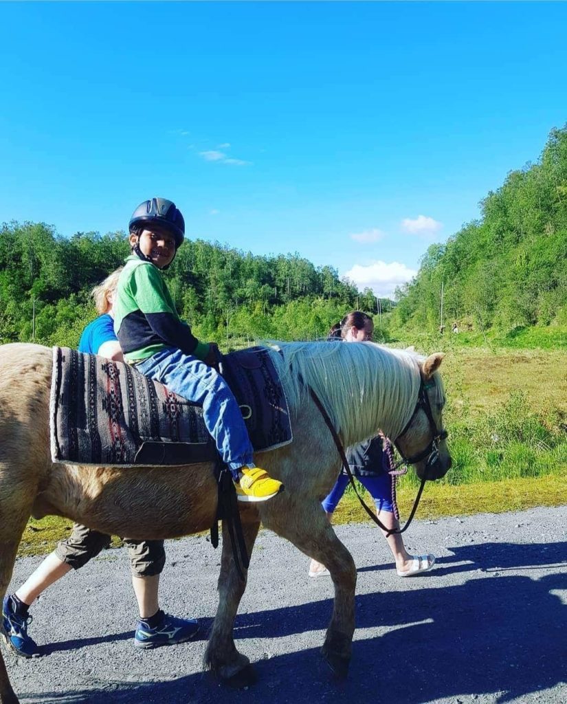 Marley rir på en hest langs veien. I bakgrunnen synes grønt gress og grønne trær. Han har på seg en grønn genser, blå bukser og gule sko. Han smiler til kamera. To assistenter går ved siden av.