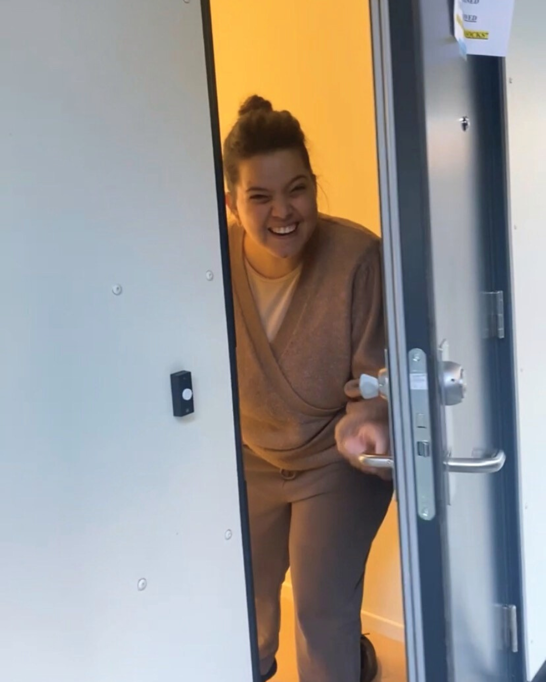 Tamarin, en ung kvinne, åpner står i inngangdøren til leiligheten sin. Hun smiler stort. Hun har bå mørk beige bukse og beige genser. Hun har brunt hår satt opp.