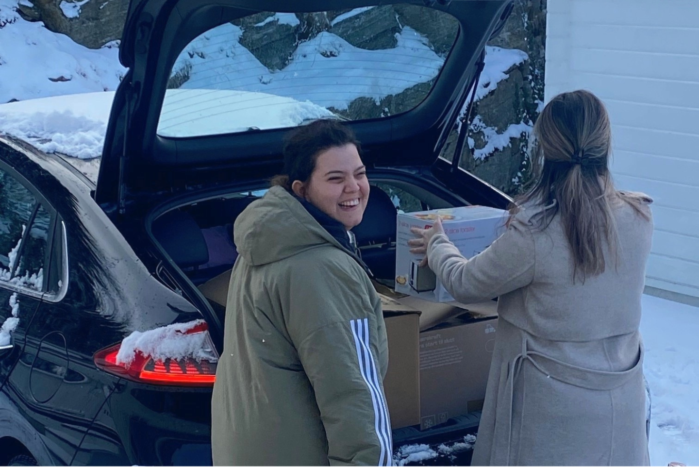 Tamarin, en ung kvinne, foran bagasjeluken på en bil sammen med kvinnelig assistent. Tamarin smiler mot kamera og assistenten løfter en flytteeske.